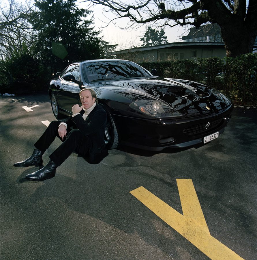 Günther Netzer sitting, leaning against black Ferrari on street.