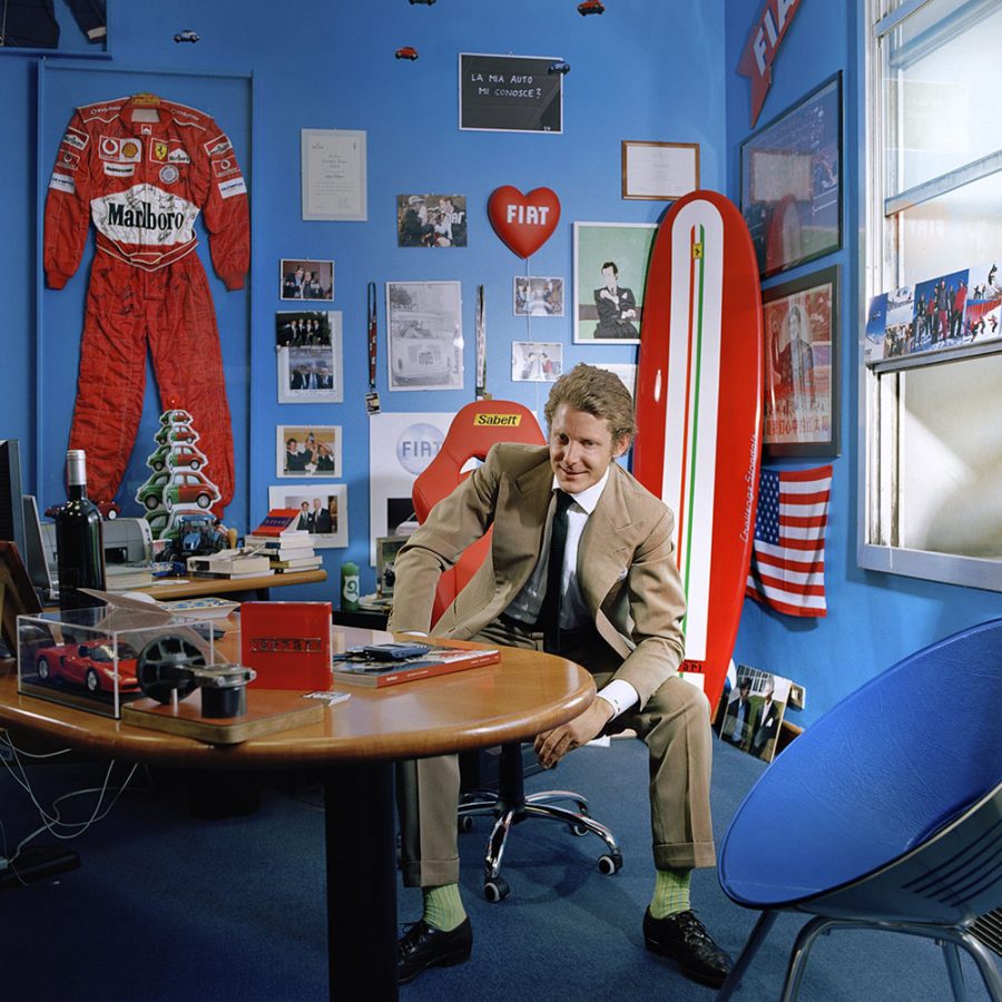 Lapo Elkann in a beige suit sitting in a blue room full of Fiat memorabilia.