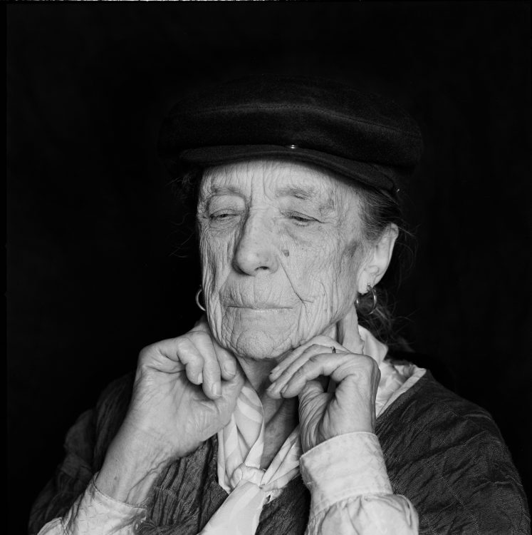 Louise Bourgeois holding hands up to her throat against a deep black background.