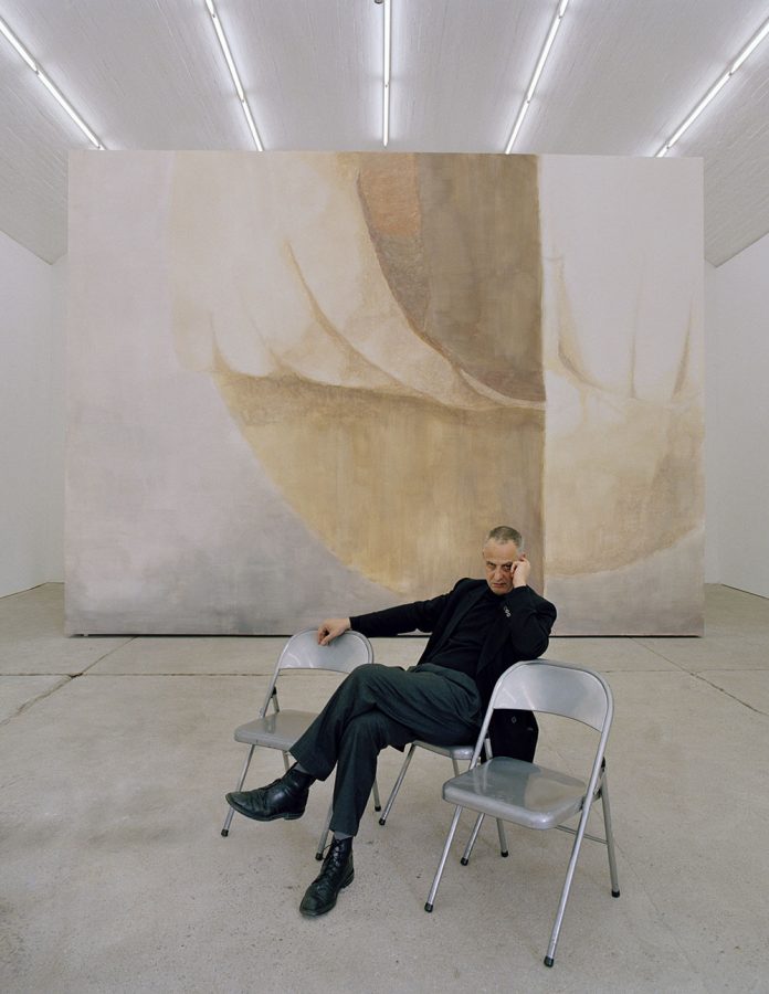Luc Tuymans is sitting in front of a beige painting that is being displayed in a large gallery.