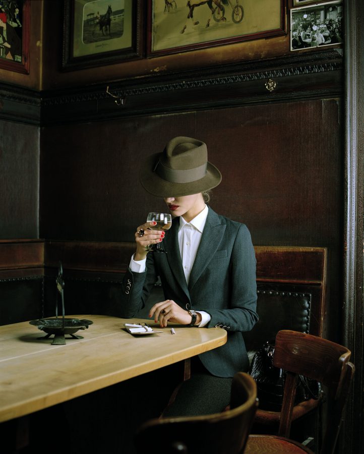 Woman in an elegant suit wearing a green fedora sits on a bar table with a wine glass in her hand.