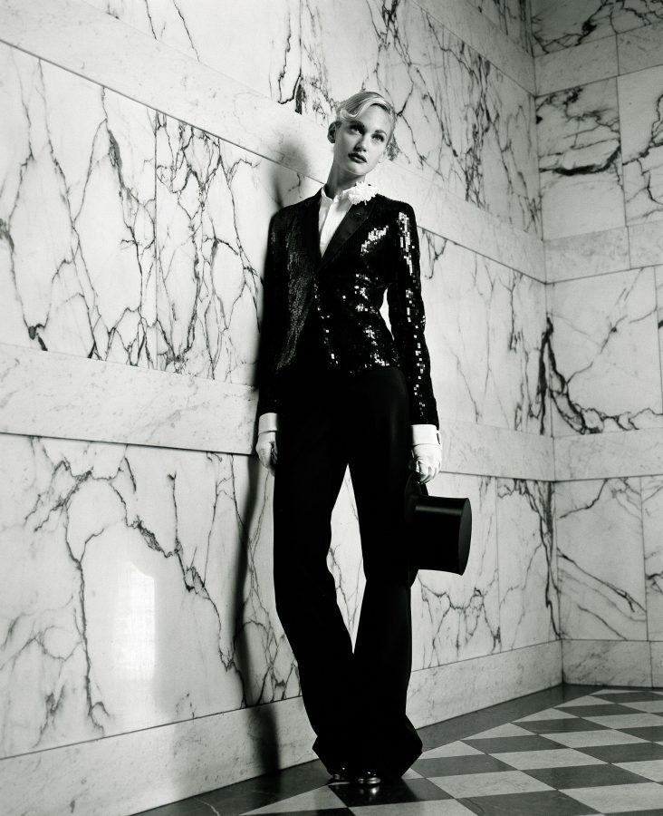 Blonde woman in a shiny black blazer leans on a white marble wall.