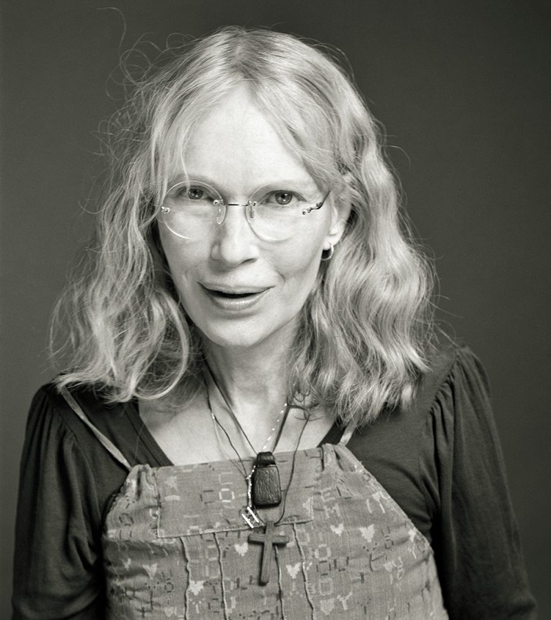 Mia Farrow wearing glasses and a wooden cross necklace.
