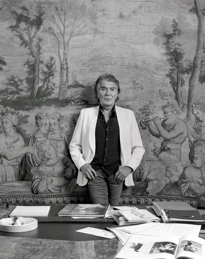 Roberto Cavalli in a white jacket a black shirt leaning on a wall behind his desk.