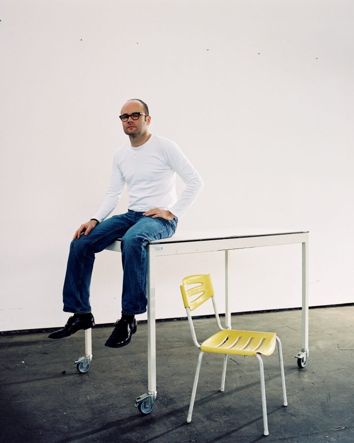 Portrait of the artist Thomas Demand in blue jeans seated on a table with a yellow chair next to it.