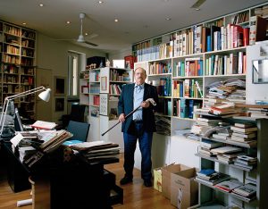 Novelist Umberto Eco holding a walking stick in his private modern library full of colorful books.