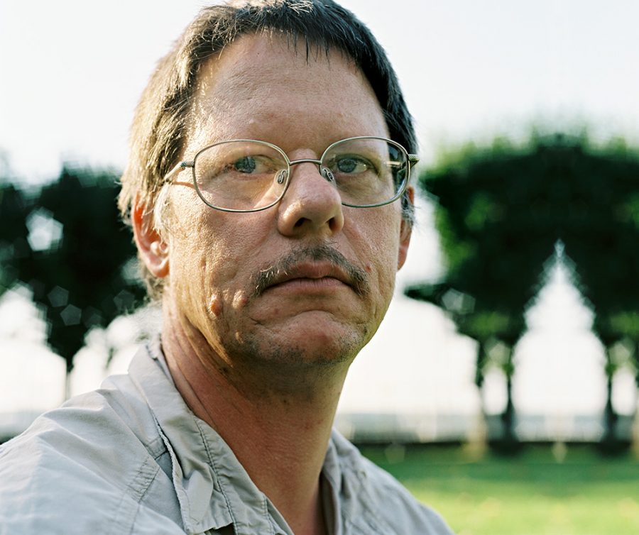 William T. Vollmann wearing glasses with green trees visible in a blurred background.