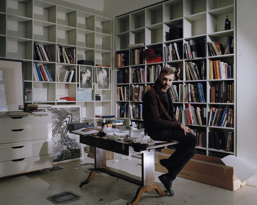 Adrian Ghenie sitting on a wooden table in his library with white bookshelves.