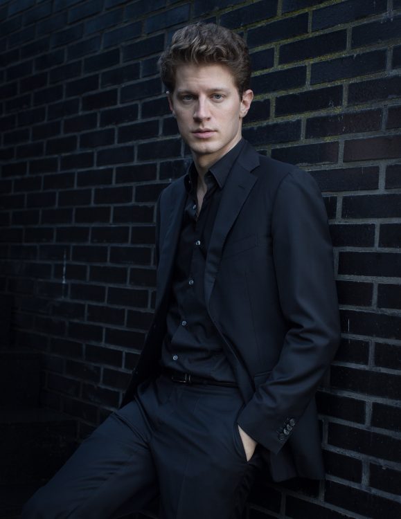 German pianist Alexander Krichel leaning on a brick wall in a black suit and black shirt.