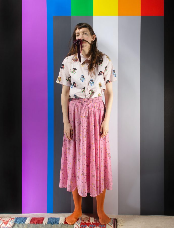 German artist Aline Schwibbe in a pink skirt standing in front of a multicolored TV test card pattern.