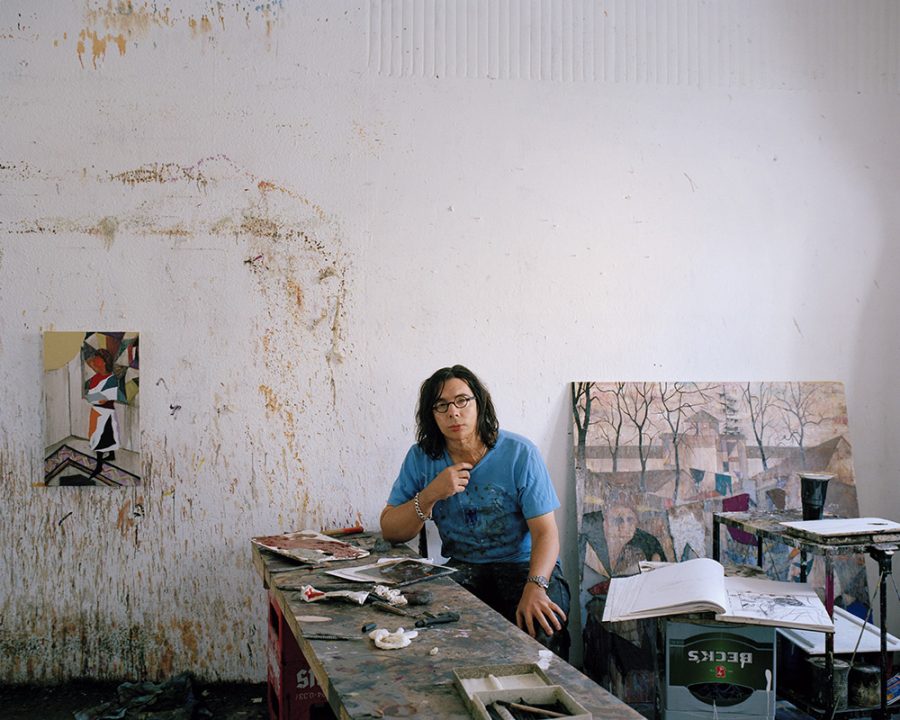 German artist Armin Boehm poses sitting in front of an improvised beer bench in front of wall covered with paint stains and two pictures behind him.
