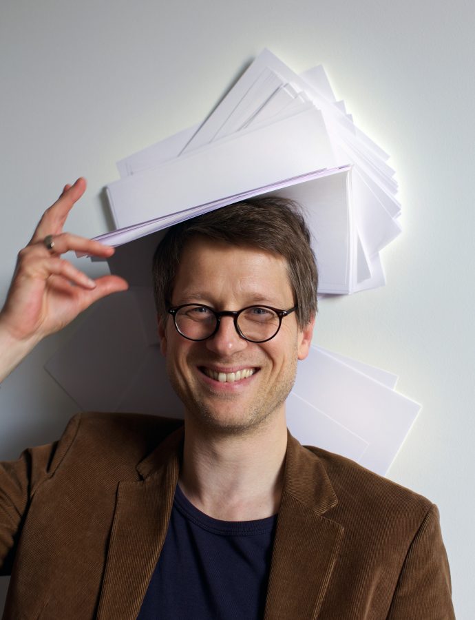 Jan Wagner with round glasses is smiling with a stack of paper on his head.