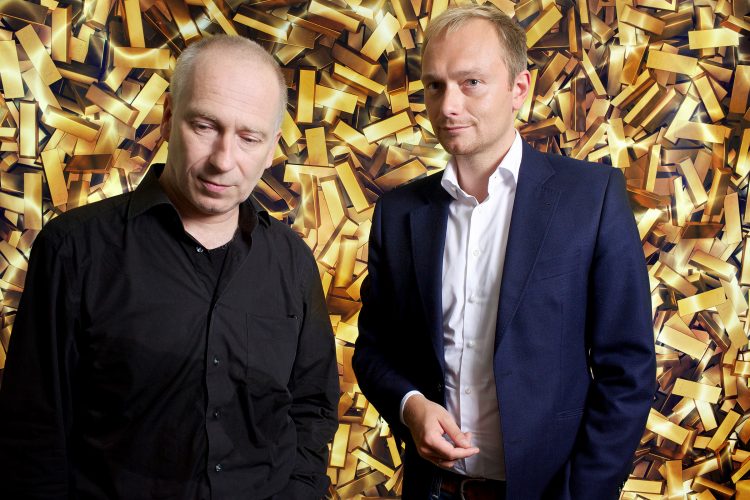 Joseph Vogl and Christian Lindner standing in front of a poster full of gold bars.