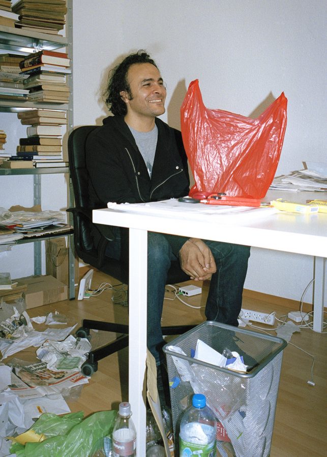 French artist Kader Attia is smiling in a messy room with a red plastic bag on a table.