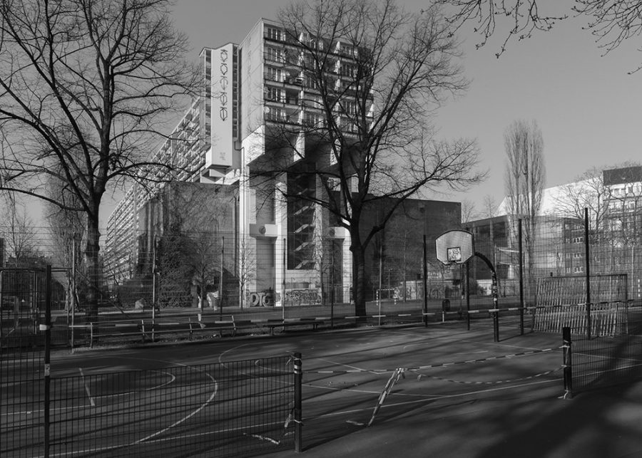 Fenced-off basketball court in front of leafless trees and a city block.