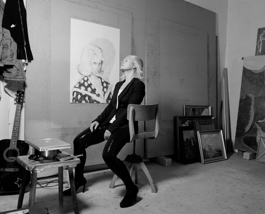 Laura Bruce sitting in her artist studio with numerous drawings and a guitar.
