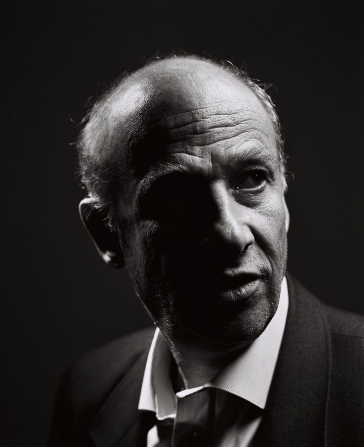 Black and white close-up portrait of Luc Bondy wearing a suit against a black background.