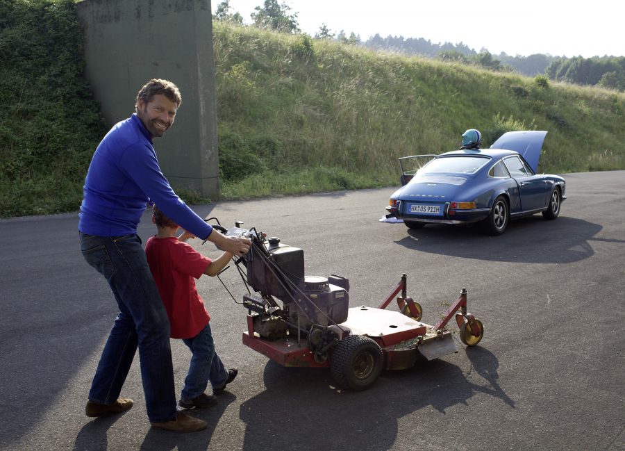 Marcus Graf von Oeynhausen-Sierstorpff and his son puhing a lawnmower with a blue Porsche in the background.