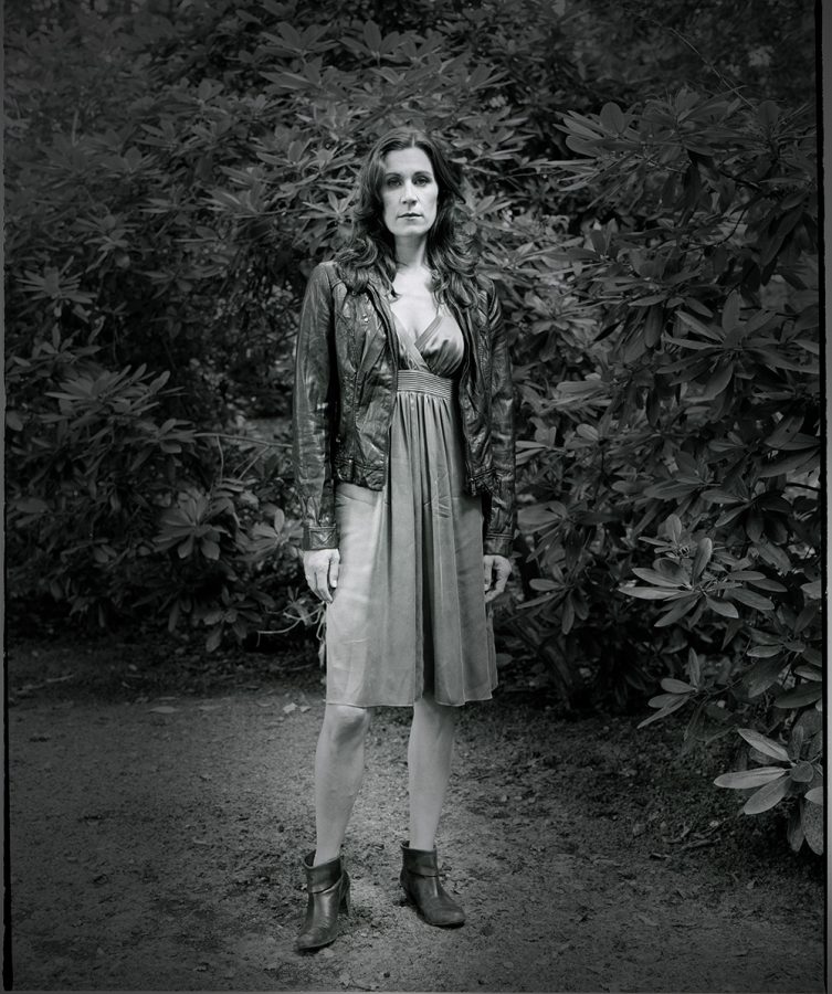 Nina Kunzendorf wearing jacket over a dress between trees and bushes.