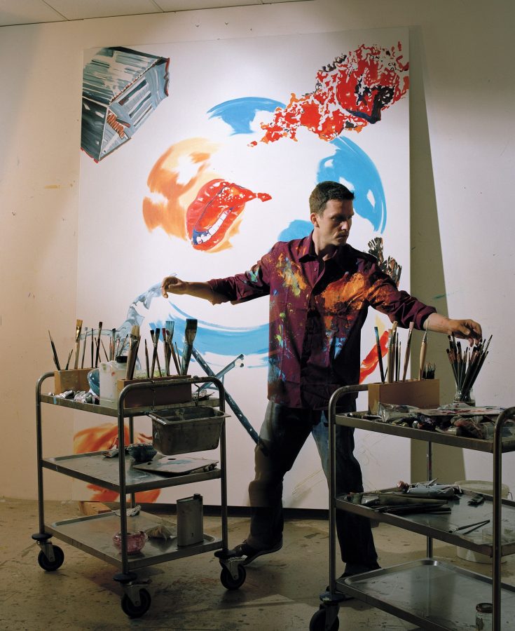 Artist Norbert Bisky in his studio in the process of painting a picture, with his painting tools on a serving cart.