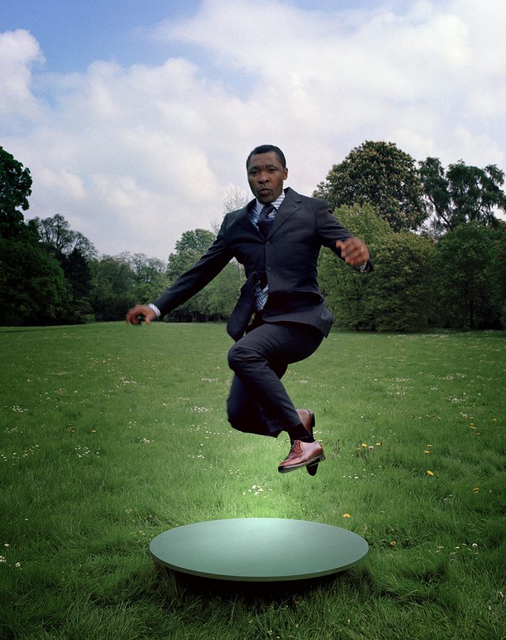 Curator Okwui Enwezor in a suit midjump on a green meadow.