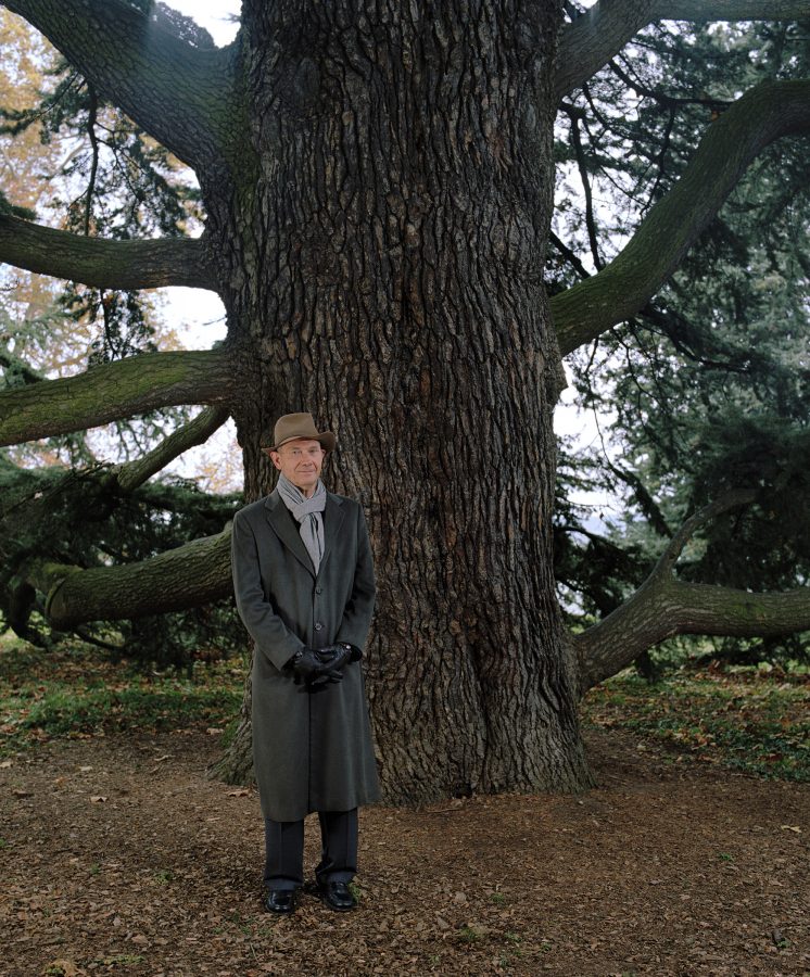 French politician Pascal Lamy in a grey coat with brown hat standing in front of a wide tree trunk.