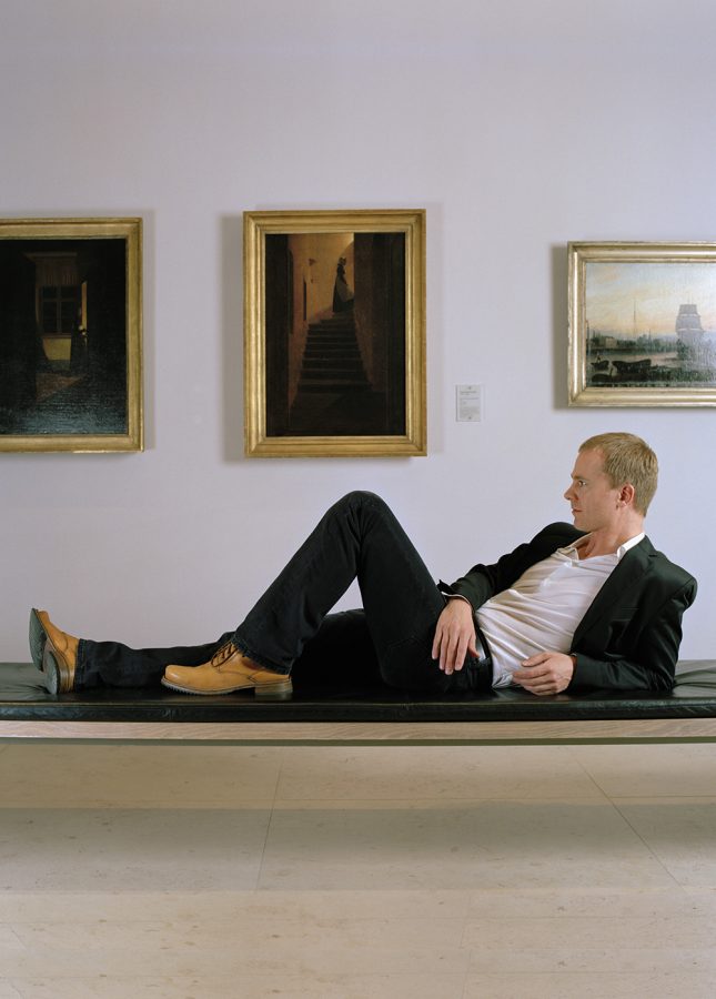 Philipp Demandt lying on a museum bench. The Caspar David Friedrich painting „Zum Licht hinaufsteigende Frau“ is hanging on a wall behind him.