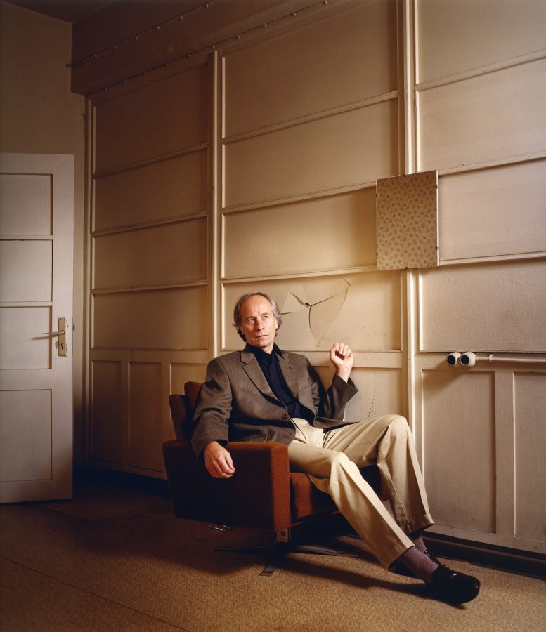 Richard Ford sitting on a couch holding his fist up with a punched-in wall behind him.