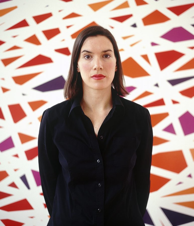 Sarah Morris wearing a black shirt with one of her geometric modernist paintings in the background.