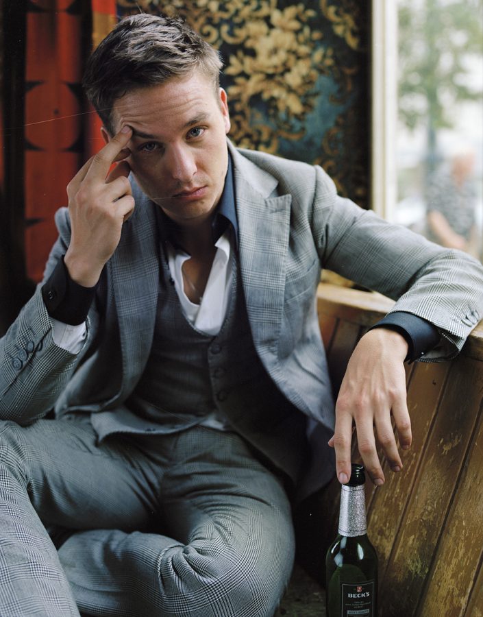 Tom Schilling in a grey three piece suit sitting on a bench with a beer.