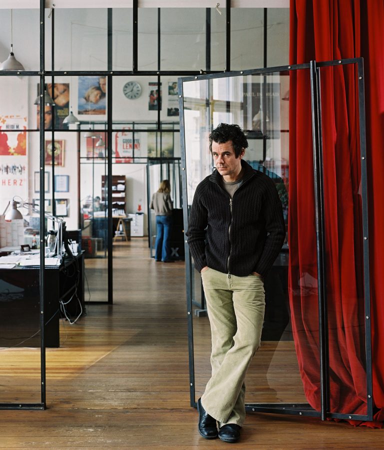 Tom Tykwer standing in a modern, transparent office loft with a red curtain next to him.