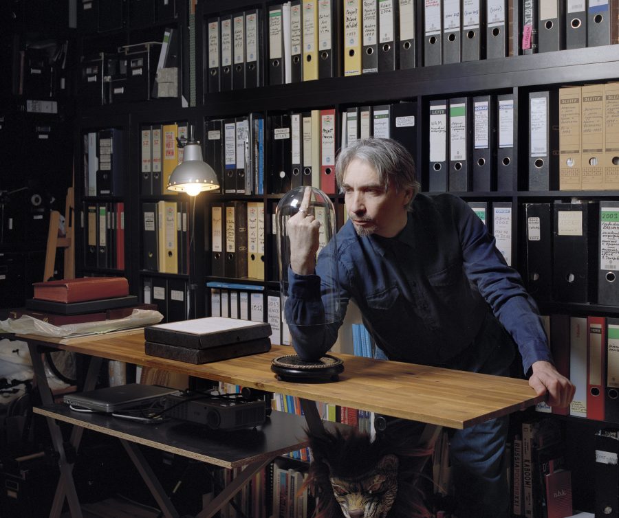 Portrait of artist Vadim Zakharov in front of his archive with a bell jar on his finger.